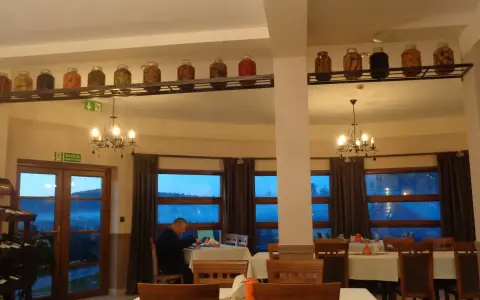 Wnętrze restauracji z oknami wychodzącymi na góry, stoły nakryte obrusami, a na półce nad nimi słoiki z przetworami.