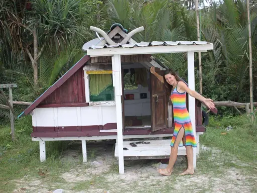 Kobieta w tęczowej sukience stoi obok małego, drewnianego domku na plaży.