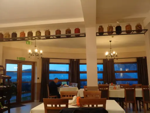 Wnętrze restauracji z oknami wychodzącymi na góry, stoły nakryte obrusami, a na półce nad nimi słoiki z przetworami.